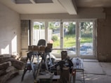 A room under renovation with construction materials scattered on the floor. There's a large window letting in sunlight and providing a view of a lush garden outside. Various tools and boxes are on a workbench, and insulation material is piled on the left.