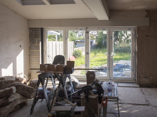 A room under renovation with construction materials scattered on the floor. There's a large window letting in sunlight and providing a view of a lush garden outside. Various tools and boxes are on a workbench, and insulation material is piled on the left.