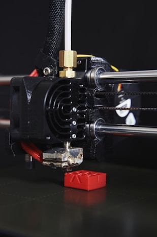 A close-up view of a 3D printer in action, with the print head creating a small red object. The printer's nozzle is prominently visible, along with metal rods and a cooling fan. The object being printed appears to have a simple geometric shape.