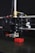 A close-up view of a 3D printer in action, with the print head creating a small red object. The printer's nozzle is prominently visible, along with metal rods and a cooling fan. The object being printed appears to have a simple geometric shape.