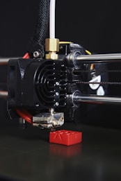 A close-up view of a 3D printer in action, with the print head creating a small red object. The printer's nozzle is prominently visible, along with metal rods and a cooling fan. The object being printed appears to have a simple geometric shape.