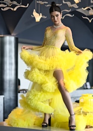 A woman is elegantly posed in a vibrant yellow ruffled gown with a slit, featuring intricate beadwork on the bodice. She accessorizes with a sparkling choker and earrings. The background includes modern décor with hanging white bird-like ornaments, set against dark-toned walls.