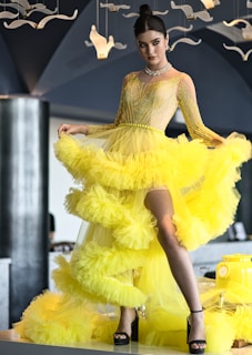 A woman is elegantly posed in a vibrant yellow ruffled gown with a slit, featuring intricate beadwork on the bodice. She accessorizes with a sparkling choker and earrings. The background includes modern décor with hanging white bird-like ornaments, set against dark-toned walls.