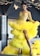A woman is elegantly posed in a vibrant yellow ruffled gown with a slit, featuring intricate beadwork on the bodice. She accessorizes with a sparkling choker and earrings. The background includes modern décor with hanging white bird-like ornaments, set against dark-toned walls.