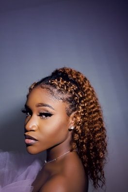 A woman with curly brown hair, styled in a high ponytail. She is wearing makeup with emphasized eyelashes and a natural lip color. Her attire includes a light, sheer fabric draped around her shoulder, and she is adorned with earrings. The lighting casts a soft glow on her face, highlighting her features against a plain backdrop.