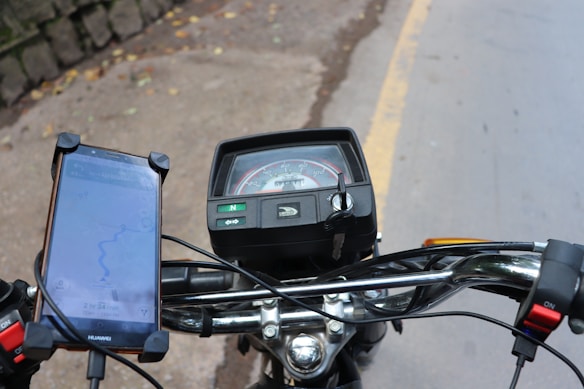 A motorcycle dashboard with a speedometer and an ignition key is visible. A smartphone mounted on the handlebars displays a GPS navigation map. The handlebars appear shiny in the light, and part of the road with a yellow line is visible in the background.
