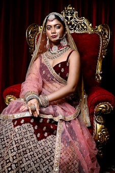A woman dressed in elaborate traditional attire sits on an ornate, gold-accented red velvet throne. Her outfit includes a richly embroidered maroon and gold lehenga, adorned with intricate jewelry, including a nose ring, earrings, necklace, and bangles. Her head is covered with a sheer dupatta, complemented by the deep red curtains in the background.