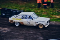 A vintage rally car with the number 39, adorned with various sponsor logos, racing on a dirt track. The vehicle appears to be moving at high speed, with motion blur visible. There are tires piled up in the background, and a group of people wearing orange jackets can be seen standing nearby on the grass.