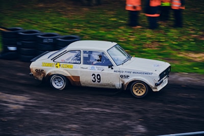 A vintage rally car with the number 39, adorned with various sponsor logos, racing on a dirt track. The vehicle appears to be moving at high speed, with motion blur visible. There are tires piled up in the background, and a group of people wearing orange jackets can be seen standing nearby on the grass.