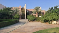 A modern residential house surrounded by lush greenery and palm trees. The building features a minimalist architectural design with clean lines and a combination of glass and concrete. The foreground consists of a stone-paved driveway and manicured lawns.