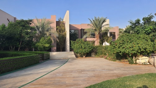 A modern residential house surrounded by lush greenery and palm trees. The building features a minimalist architectural design with clean lines and a combination of glass and concrete. The foreground consists of a stone-paved driveway and manicured lawns.