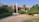 A modern residential house surrounded by lush greenery and palm trees. The building features a minimalist architectural design with clean lines and a combination of glass and concrete. The foreground consists of a stone-paved driveway and manicured lawns.