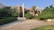 A modern residential house surrounded by lush greenery and palm trees. The building features a minimalist architectural design with clean lines and a combination of glass and concrete. The foreground consists of a stone-paved driveway and manicured lawns.