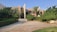A modern residential house surrounded by lush greenery and palm trees. The building features a minimalist architectural design with clean lines and a combination of glass and concrete. The foreground consists of a stone-paved driveway and manicured lawns.