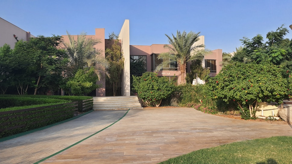 A modern residential house surrounded by lush greenery and palm trees. The building features a minimalist architectural design with clean lines and a combination of glass and concrete. The foreground consists of a stone-paved driveway and manicured lawns.