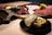 A luxurious and artistic food presentation featuring a black plate with a decorative, cream-colored bowl that is partially open to reveal gold leaf. The background displays a wooden serving board with slices of raw meat and garnishes, including flowers and herbs. The atmosphere is elegant and sophisticated.