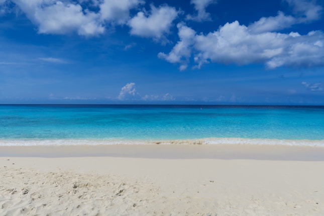 A serene beach scene with clear turquoise waters meeting a sandy shore under a vibrant blue sky scattered with fluffy white clouds.