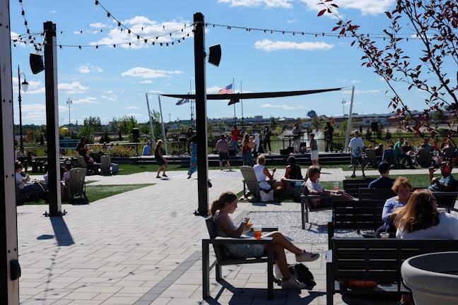 People are gathered in an outdoor space with seating areas, enjoying a sunny day. A stage is set up with performers, and there are string lights overhead. Trees and a few flags can be seen in the background, creating a lively and relaxed atmosphere.