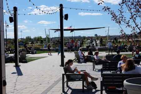 People are gathered in an outdoor space with seating areas, enjoying a sunny day. A stage is set up with performers, and there are string lights overhead. Trees and a few flags can be seen in the background, creating a lively and relaxed atmosphere.