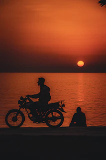Swavoy sharing a moment while riding a motorcycle along a coastal highway at sunset.