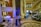 A luxurious interior space with a grand staircase and ornate decor. A small musical ensemble, including a violinist and a singer, performs near a white grand piano. Several people are seated, watching the performance, while others casually walk by. The setting appears elegant and relaxed.