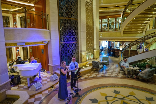 A luxurious interior space with a grand staircase and ornate decor. A small musical ensemble, including a violinist and a singer, performs near a white grand piano. Several people are seated, watching the performance, while others casually walk by. The setting appears elegant and relaxed.