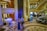 A luxurious interior space with a grand staircase and ornate decor. A small musical ensemble, including a violinist and a singer, performs near a white grand piano. Several people are seated, watching the performance, while others casually walk by. The setting appears elegant and relaxed.