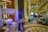 A luxurious interior space with a grand staircase and ornate decor. A small musical ensemble, including a violinist and a singer, performs near a white grand piano. Several people are seated, watching the performance, while others casually walk by. The setting appears elegant and relaxed.