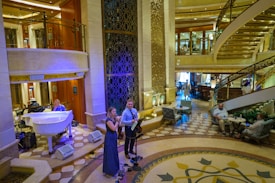 A luxurious interior space with a grand staircase and ornate decor. A small musical ensemble, including a violinist and a singer, performs near a white grand piano. Several people are seated, watching the performance, while others casually walk by. The setting appears elegant and relaxed.