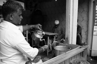 A man in a white shirt is pouring liquid from a teapot into a cup, with a small shop setting in the background. Another man is seated at a table, wearing traditional clothing and a cap, seemingly engaged in a quiet activity. The environment suggests a casual street vendor or tea stall atmosphere, with visible kitchen utensils and vessels.