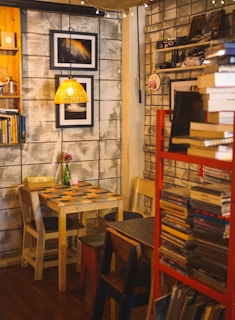 A cozy cafe interior with a small wooden table and two mismatched chairs. A woven lampshade hangs above the table, casting a warm light. The walls are adorned with framed artwork, books, and small decorative items. A red shelf on the right side is packed with books and records, adding to the vintage atmosphere.
