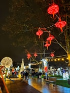 A festive street scene is illuminated by bright lights at night. Red lanterns hang from bare tree branches, and a variety of decorative lights are strung across the area. A group of people, some with children, walk along the wet pavement. Elaborate light displays include a tall, illuminated Christmas tree and various shaped lights on the grass.