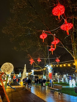A festive street scene is illuminated by bright lights at night. Red lanterns hang from bare tree branches, and a variety of decorative lights are strung across the area. A group of people, some with children, walk along the wet pavement. Elaborate light displays include a tall, illuminated Christmas tree and various shaped lights on the grass.