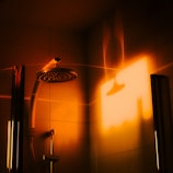 A shower head and its mount inside a shower cubicle, illuminated by a warm, orange light. The light casts a shadow that reflects on the glass and tiles, creating a cozy and inviting atmosphere.