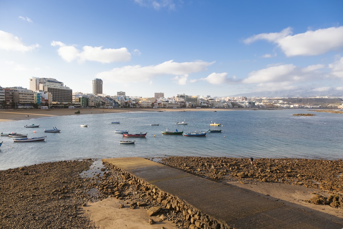 A bay in Las Palmas with small boats on the water, rocks and sand in the foreground and the white line of the city along the shore