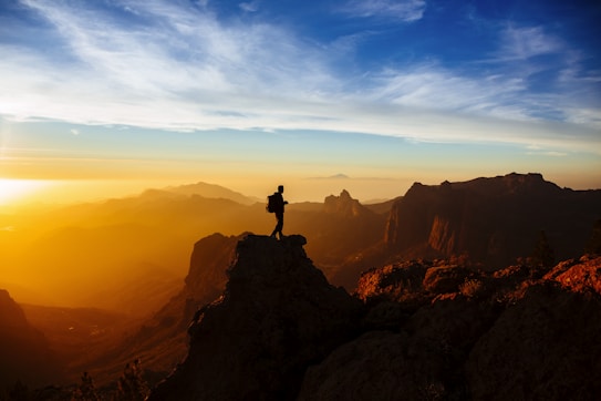 A lone hiker stands atop a rocky peak, silhouetted against a vibrant sunset backdrop. The landscape features rolling hills and distant mountains fading into the horizon. The sky is a mix of deep blues and warm orange hues, creating a dramatic contrast between the cool sky and the warm, glowing atmosphere.