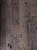 A close-up view of an old, worn-out wooden floor with numerous dark spots and blemishes. The planks show signs of aging and wear, including scratches, dents, and some areas where they appear to be splintering.