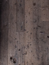 A close-up view of an old, worn-out wooden floor with numerous dark spots and blemishes. The planks show signs of aging and wear, including scratches, dents, and some areas where they appear to be splintering.