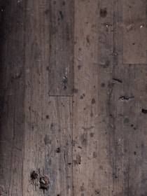 A close-up view of an old, worn-out wooden floor with numerous dark spots and blemishes. The planks show signs of aging and wear, including scratches, dents, and some areas where they appear to be splintering.