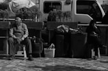 Two people sit on stools surrounded by bags and luggage on a tiled area. A white vehicle is parked behind them, and potted plants line a low wall parallel to the vehicle. One person appears contemplative while the other seems relaxed.