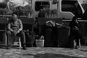 Two people sit on stools surrounded by bags and luggage on a tiled area. A white vehicle is parked behind them, and potted plants line a low wall parallel to the vehicle. One person appears contemplative while the other seems relaxed.
