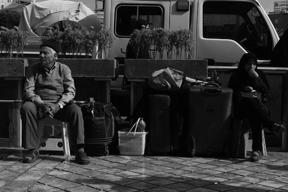 Two people sit on stools surrounded by bags and luggage on a tiled area. A white vehicle is parked behind them, and potted plants line a low wall parallel to the vehicle. One person appears contemplative while the other seems relaxed.
