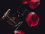 A dark and moody composition featuring a glossy black perfume bottle with elegant golden text. Water droplets add a fresh and luxurious feel. Surrounding it are deep red rose petals, enhancing the romantic and sophisticated atmosphere.