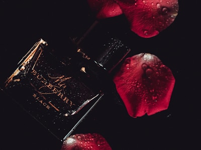 A dark and moody composition featuring a glossy black perfume bottle with elegant golden text. Water droplets add a fresh and luxurious feel. Surrounding it are deep red rose petals, enhancing the romantic and sophisticated atmosphere.