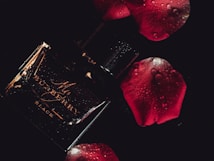 A dark and moody composition featuring a glossy black perfume bottle with elegant golden text. Water droplets add a fresh and luxurious feel. Surrounding it are deep red rose petals, enhancing the romantic and sophisticated atmosphere.