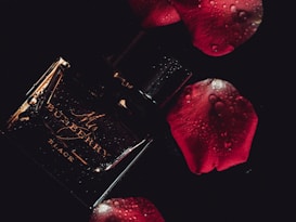 A dark and moody composition featuring a glossy black perfume bottle with elegant golden text. Water droplets add a fresh and luxurious feel. Surrounding it are deep red rose petals, enhancing the romantic and sophisticated atmosphere.