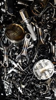 A chaotic assortment of various metal engine parts, including pistons, connecting rods, nuts, bolts, and springs. The components have a metallic sheen, and some show signs of wear and oxidation, suggesting they are used or discarded.