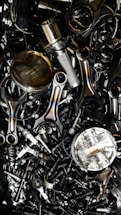 A chaotic assortment of various metal engine parts, including pistons, connecting rods, nuts, bolts, and springs. The components have a metallic sheen, and some show signs of wear and oxidation, suggesting they are used or discarded.