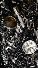 A chaotic assortment of various metal engine parts, including pistons, connecting rods, nuts, bolts, and springs. The components have a metallic sheen, and some show signs of wear and oxidation, suggesting they are used or discarded.