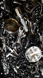 A chaotic assortment of various metal engine parts, including pistons, connecting rods, nuts, bolts, and springs. The components have a metallic sheen, and some show signs of wear and oxidation, suggesting they are used or discarded.
